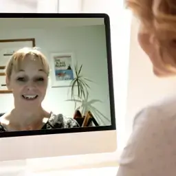 A woman smiling on a video call displayed on a computer screen while another person watches from behind.
