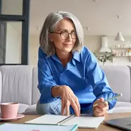 A mature woman with gray hair and glasses, wearing a blue shirt, sits on a couch with a notebook and pen, smiling while studying at home.